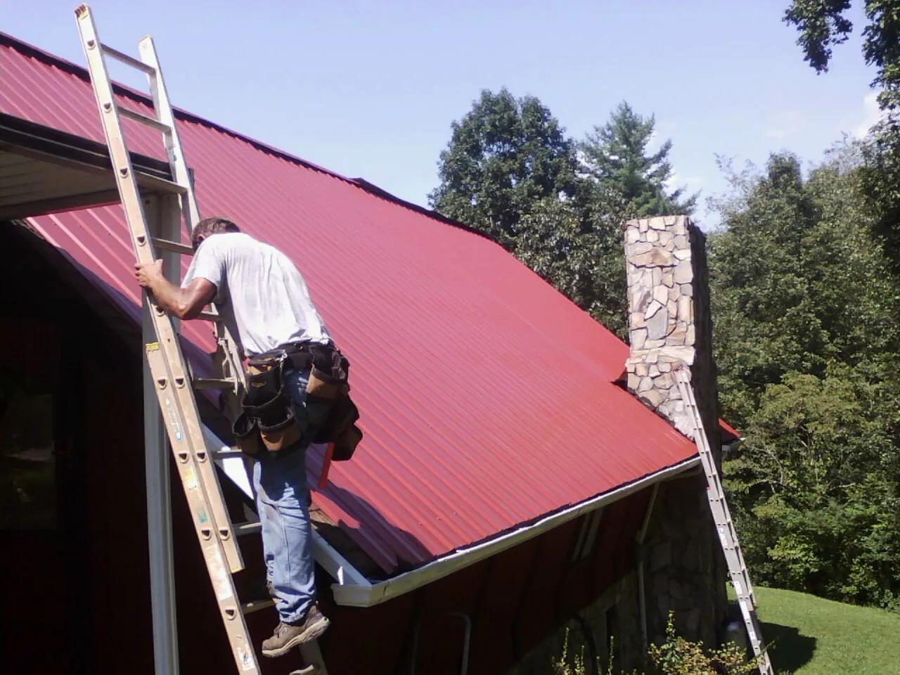 Roofer on a ladder beside a red metal roof and stone chimney — Soffit & Fascia Repair by Roofing Brothers in Eagle Rock